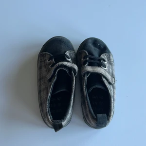 Rutiga sneakers i svart och grått - Snygga sneakers i svart och grått med rutigt mönster. Skorna har snörning framtill och en praktisk kardborreknäppning. Ovandelen är i textil och sulan är platt och svart. Perfekta för en cool och avslappnad stil.