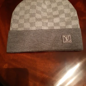 Grå rutig mössa från Louis Vuitton - Snygg grå mössa från Louis Vuitton med klassiskt rutigt mönster och uppvikt kant. LV-logga framtill och diskret design som passar perfekt till streetwear. Tillverkad i mjukt material för skön känsla och stilren look.