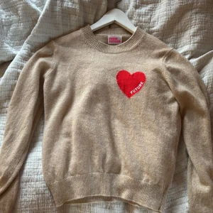 Beige kashmirtröja From Future med hjärta - Supermjuk beige kashmirtröja från From Future med ett rött hjärta och texten 'FUTURE' broderat på bröstet. Rund halsringning, ribbade muddar och långärmad modell. Perfekt för dig som vill ha en mysig och snygg statementtröja.
