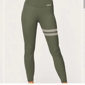 Olivgröna leggings från Stronger - Snygga olivgröna leggings från Stronger med två vita ränder på ena låret. Hög midja och tight passform, perfekta för träning. Tillverkade i stretchigt syntetmaterial som andas. Diskret logga framtill och meshdetaljer på vaderna för extra ventilation. Dessa säljs även inte på jemsidan längre! Nypris - 700