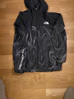 Svart vindjacka från The North Face - Säljer en svart vindjacka från The North Face med vit topografiskt mönster och logga på bröstet och ärmen. Jackan har huva, dragkedja framtill och är tillverkad i lätt polyester. Perfekt för blåsiga dagar och snygg streetstil.