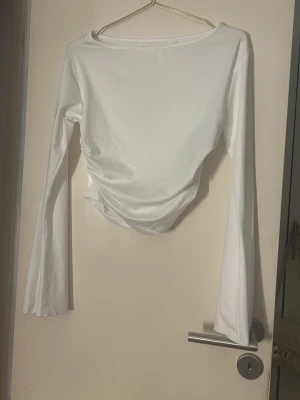 Vit långärmad topp med drapering - Säljer en stilren vit långärmad topp med lätt drapering i sidan. Toppen har en enkel, rund halsringning och är tillverkad i ett mjukt, stretchigt material som sitter snyggt på kroppen. Perfekt att matcha med jeans eller kjol för en clean look. Aldrig använd. Stl L men liten i storlek