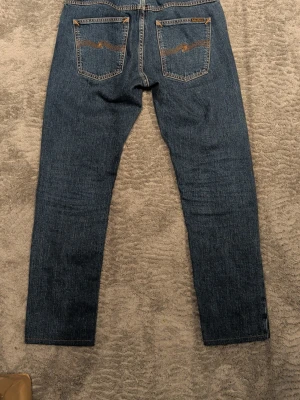 Blå jeans från Nudie Jeans Co - Säljer ett par klassiska blå jeans från Nudie Jeans Co med raka ben och snygga kontrastsömmar. Jeansen har ikoniska bakfickor med vågig söm och patch i läder bak i midjan. Tillverkade i bomull och har en tidlös look som passar till allt.