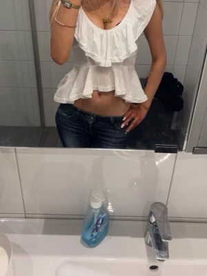 Vit volangblus med öppen rygg - Säljer en superfin vit blus med volanger och bred urringning. Toppen är croppad och har en snygg öppen rygg med knytband. Perfekt till jeans eller kjol för en fräsch look. Materialet känns lätt och luftigt, perfekt för varmare dagar.