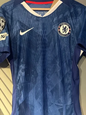 Chelsea Nike matchtröja blå - Säljer en officiell Chelsea matchtröja från Nike i marinblått med vita detaljer och klubbmärke på bröstet. Tröjan har ett subtilt mönster i tyget och patch på ärmen. Perfekt för dig som älskar fotboll och vill stötta ditt lag.