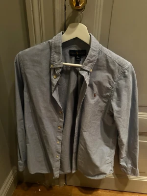 Ljusblå skjorta från Ralph Lauren - Klassisk ljusblå skjorta från Ralph Lauren med button-down krage och broderad logga på bröstet. Skjortan har långa ärmar och stängs med knappar framtill. Perfekt för en clean och stilren look.