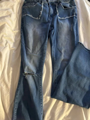 Blå jeans med slitningar och raka ben - Säljer ett par blå jeans med raka ben och slitningar vid knät samt fransiga detaljer runt fickorna. Jeansen har klassisk femficksdesign och är tillverkade i ett mjukt denimtyg. Perfekta för en avslappnad och trendig look.