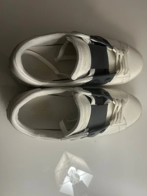 Valentino Garavani Open sneakers vit/mörkblå - Säljer ett par Valentino Garavani Open sneakers i vitt skinn med bred svart läderdetalj över snörningen. Klassisk rund tå och platt sula. Kommer med originalpåse och box. Perfekta för dig som vill ha en clean men lyxig look.