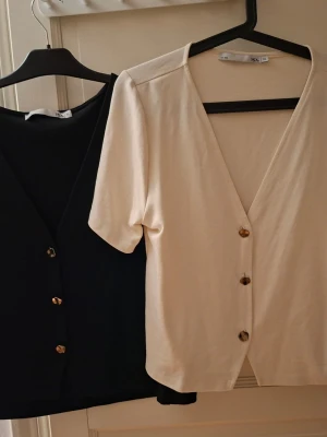 Två kortärmade blusar från 157 - Säljer två stilrena blusar från 157 i storlek XL. En är svart och en är beige, båda med v-ringning och snygga knappar framtill. De har korta ärmar och är gjorda i mjuk bomull, perfekta för en clean och enkel look.  100kr st 