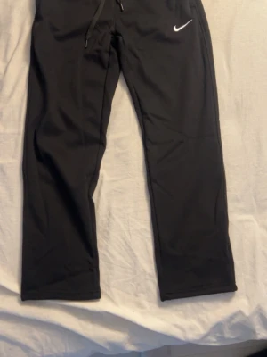Svarta mjukisbyxor från Nike M - Svarta mjukisbyxor från Nike med klassisk vit logga på låret. Byxorna har resår i midjan med snörning och raka ben. Perfekta för chill eller träning, tillverkade i mjukt material som känns skönt mot huden. Perfekt till gym eller bara chill