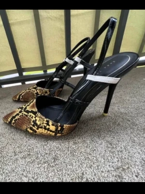 Ormmönstrade pumps med hög klack - Säljer ett par trendiga pumps från Zara med ormmönstrad tå i bruna och gula toner. Skorna har spetsig tå, hög klack och svarta samt vita remmar i glansigt material. Perfekta för dig som vill sticka ut med en edgy look.