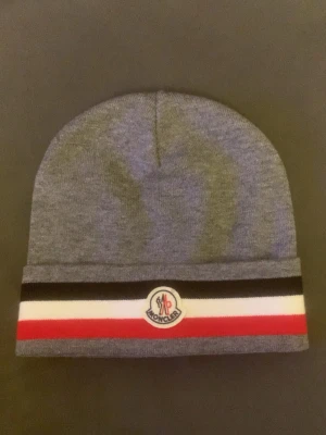 Grå Moncler mössa med ränder - Säljer en grå Moncler mössa med klassiska ränder i svart, vitt och rött längst ner. Mössan har Moncler-logga framtill och är tillverkad i mjukt material som känns skönt mot huvudet. Perfekt accessoar för dig som vill ha en clean och stilren look.