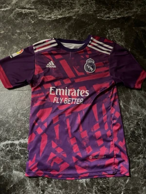  Real Madrid Benzema matchtröja - Säljer en lila och rosa Real Madrid matchtröja från Adidas med Benzema och nummer 9 på ryggen. Tröjan har ett grafiskt mönster, vita Adidas-ränder på axlarna och klubbmärke på bröstet. Tillverkad i lätt polyester med Aeroready-teknologi.