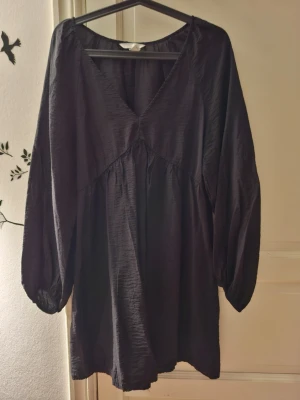 Svart kortklänning med v-ringning - Säljer en svart kortklänning med v-ringning och långa, luftiga ärmar. Klänningen har en lös passform och är tillverkad i ett lätt, något glansigt material som ger en snygg struktur. Perfekt för dig som gillar en enkel men stilren look.