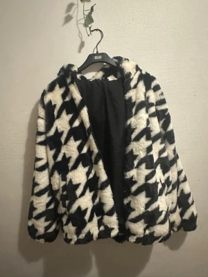 Svartvit pälsjacka från H&M Divided - Fluffig och mjuk pälsjacka från H&M Divided i storlek L. Jackan har ett svartvitt rutigt mönster med stora grafiska former och är helfodrad. Perfekt för dig som vill sticka ut med en cool och trendig look. Jackan har huva och långa ärmar.