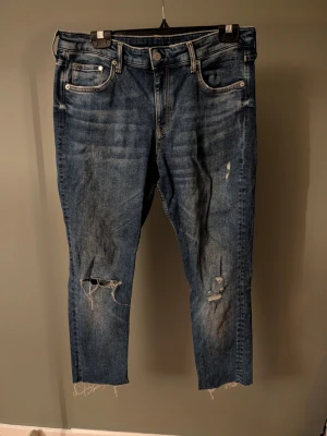Blå croppade jeans med slitningar - Säljer ett par blå croppade jeans med råa, fransiga benslut och slitningar på både knän och lår. Jeansen har klassisk femficksmodell och är tillverkade i denim med en rak passform. Perfekta för en avslappnad och trendig look.