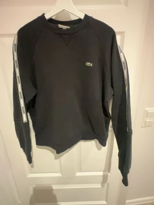 Svart sweatshirt från Lacoste L - Svart sweatshirt från Lacoste i relaxed fit med klassisk krokodil-logga på bröstet och vita band med Lacoste-tryck längs ärmarna. Tröjan har långa ärmar och rund hals. Perfekt för en chill och sportig look.