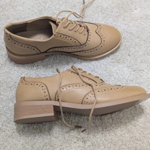 Beige brogues med snörning - Riktigt bekväma snöskor med dämpad sula, oanvända 🍓                            Beige brogues med klassiska perforerade detaljer och snörning. Skorna har låg klack och platt sula, tillverkade i syntetmaterial med rund tå. Perfekta för dig som vill ha en stilren och tidlös look.