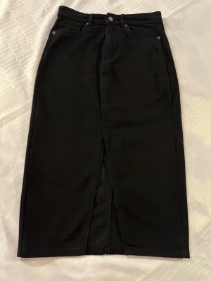 Svart långkjol från ONLY i denim M  - Säljer en svart långkjol från ONLY i denim med klassisk femficksdesign, bälteshällor och en snygg slits framtill. Kjolen har knapp och dragkedja i midjan och är perfekt för dig som gillar en stilren och enkel look.