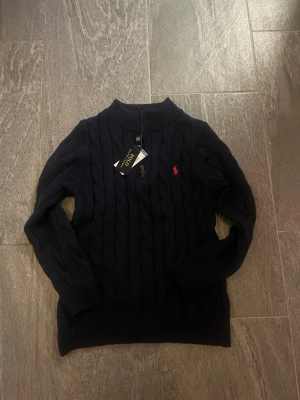 Mörkblå stickad tröja Polo Ralph Lauren - Säljer en mörkblå stickad tröja från Polo Ralph Lauren i storlek S. Tröjan har kabelstickat mönster, ribbad krage och en half zip-dragkedja vid halsen. Klassisk röd logga broderad på bröstet. Perfekt för dig som gillar stilrena och tidlösa plagg.