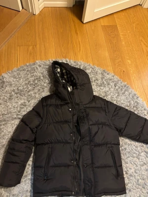 Svart pufferjacka från Burberry - Säljer en svart pufferjacka från Burberry med stor huva och rutigt innerfoder i svart, vitt och rött. Jackan har dragkedja och knappar framtill samt två fickor med dragkedja. Klassisk Burberry-patch på ärmen och riktigt skön passform för kalla dagar. 