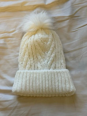 Vit stickad mössa med fuskpälsboll - Supermysig vit stickad mössa från Newbie med bred uppvikt kant och en fluffig fuskpälsboll på toppen. Perfekt för kalla dagar och ger en söt vinterlook. Klassisk kabelstickning och mjukt material som håller dig varm.