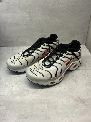 Nike TN air vita och svarta - Nike TN air sneakers i vitt mesh med svarta vågiga detaljer, röd Swoosh och blå sula med gula och röda inslag. Skorna har klassisk Tn-logga bak och synlig Air-enhet i sulan. Svart snörning och vadderad plös för extra komfort.