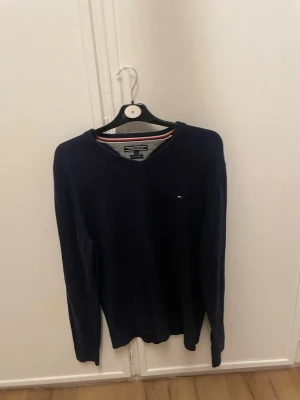 Mörkblå tröja Tommy Hilfiger L - Snygg mörkblå tröja från Tommy Hilfiger i storlek L. Tillverkad i en mjuk mix av pima bomull och kashmir. Klassisk v-ringning och diskret logga broderad på bröstet. Perfekt till jeans eller chinos för en clean look.