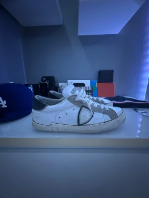 Philippe Model vita sneakers med grå detaljer - Säljer ett par Philippe Model sneakers i vitt skinn med grå mockadetaljer och svart logotyp på sidan. Klassisk låg modell med snörning och platt sula. Skorna har en cool, urban vibe och passar perfekt till streetwear eller casual outfits.