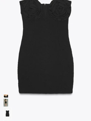 Svart kort klänning med blommor - Supersnygg svart kort klänning med hjärtformad urringning och stora blommor på bysten. Axelbandslös modell som sitter tight och framhäver figuren. Perfekt för dig som vill sticka ut med en unik detalj upptill. Helt ny med prislapp på!