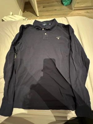 Mörkblå långärmad henley från Gant - Säljer en mörkblå långärmad henley från Gant i regular fit. Skjortan har klassisk krage, två knappar framtill och diskret Gant-logga broderad på bröstet. Tillverkad i mjuk bomull som känns skön mot huden.