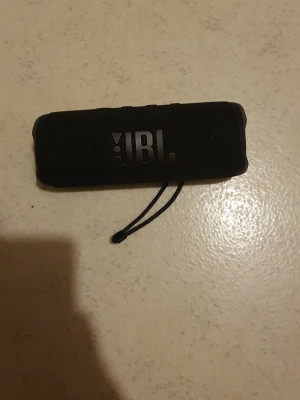 JBL Flip 6 Bluetooth-högtalare - JBL Flip 6 är en portabel Bluetooth-högtal, känd för kraftfullt ljud och robust design. Svart färg, med tydlig JBL-logga och handledsrem. Högtalaren ser ut att vara i gott skick utan synliga skador eller slitage. Perfekt för musik hemma eller utomhus.