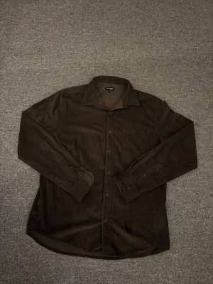 Brun manchesterskjorta On Our Terms M - Säljer en brun manchesterskjorta från On Our Terms i storlek M. Skjortan har klassisk krage, bröstficka och knäppning framtill. Långärmad modell med mjuk känsla och diskret struktur i tyget. Perfekt för dig som gillar stilrena plagg med lite retrovibe.