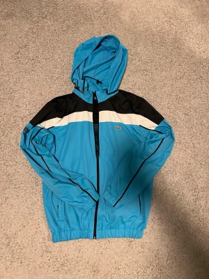Blå vindjacka från Lacoste Sport - Säljer en blå vindjacka från Lacoste Sport med svart och vit panel upptill, huva och dragkedja framtill. Jackan har elastiska muddar, två fickor och klassisk Lacoste-logga på bröstet. Perfekt för dig som vill ha en sportig och clean look.