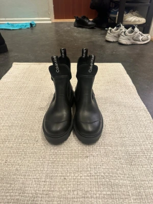Svarta chelsea boots från Calvin Klein - Säljer ett par svarta chelsea boots från Calvin Klein Jeans. Skorna har grov sula, elastiska paneler på sidorna och dragflikar med logga både fram och bak. Tillverkade i skinnliknande syntetmaterial och har en chunky, modern look.