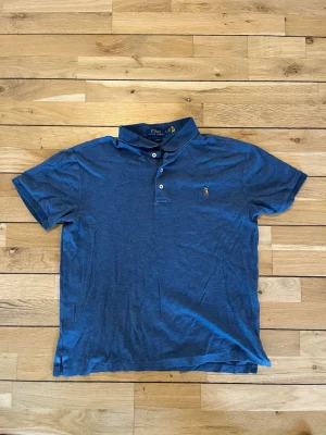 Blå pikétröja från Polo Ralph Lauren - Säljer en klassisk blå pikétröja från Polo Ralph Lauren i modellen Slim Fit. Tröjan har korta ärmar, krage och två knappar framtill. Den ikoniska broderade loggan syns på bröstet. Perfekt till jeans eller shorts för en clean look.