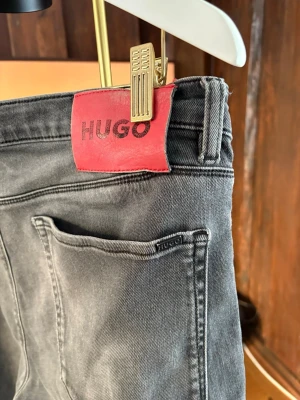 Grå jeans från Hugo 1708 - Snygga grå jeans från Hugo med klassisk femficksdesign och röda Hugo-detaljer på etikett och knapp. Modellen är 1708 och har en slim passform med raka ben. Jeansen är tillverkade i bomull med en lätt tvättad look. 