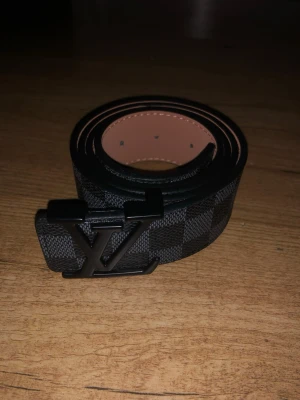 Lv bälte  - Säljer ett svart och grått LV-bälte med klassiskt rutigt mönster och stor LV-logga i metall framtill. Bältet är gjort i skinn med beige insida och har en stilren, modern look som passar till jeans eller kostymbyxor.