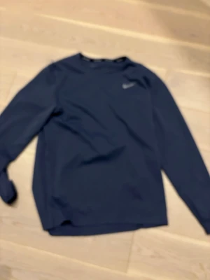 Blå Nike Dri-Fit långärmad tröja - Nike Dri-Fit långärmad tröja i marinblått med diskret Nike-logga på bröstet. Tillverkad i ett lätt och funktionellt material som andas, perfekt för löpning eller träning. Tröjan har rund hals och är designad för att transportera bort fukt.