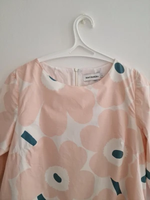 Marimekko blommig kortklänning - Supersöt a-linjeformad kortklänning från Marimekko i ljusa toner med stort blommigt mönster Unikko i rosa, vitt och mörkblått. Klänningen har trekvartsärm, rund halsringning och dragkedja bak. Tillverkad i mjuk bomull, perfekt för vår och sommar.
