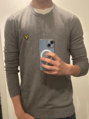 Grå stickad tröja från Lyle & Scott - Säljer en stilren grå stickad tröja från Lyle & Scott med deras klassiska gula logga på bröstet. Tröjan har rund halsringning, ribbade muddar och är långärmad. Perfekt till höst och vinter för en clean och enkel look.