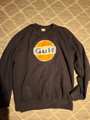 sweatshirt med Gulf-logga - Cool svart sweatshirt med stor retro Gulf-logga i orange, blått och vitt på bröstet. Klassisk rund hals och långa ärmar. Perfekt för dig som gillar vintage och streetwear vibes.