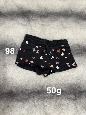 Svarta shorts med fjärilsmönster - Säljer ett par svarta shorts med elastisk midja och snörning. Shortsen har ett gulligt fjärilsmönster i vitt, rosa och silver. Perfekta för varma dagar och riktigt sköna att ha på sig.🌸 Fint använt skick. Storlek 98