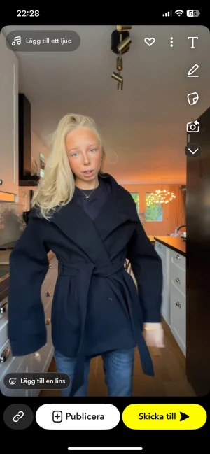 Svart omlottkappa med bälte - Stilren svart kappa med omlottmodell och brett bälte i midjan. Jackan har långa ärmar, bred krage och knäppning framtill. Perfekt för dig som gillar en clean look och vill ha något som funkar till många outfits. Storlek 158/164