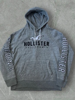 Grå hoodie från Hollister i storlek L - Snygg grå hoodie från Hollister i storlek L med stor logga och texten CALIFORNIA på bröstet och längs båda ärmarna. Tröjan har huva med snörning och en stor magficka framtill. Mjuk insida och klassisk streetstil.