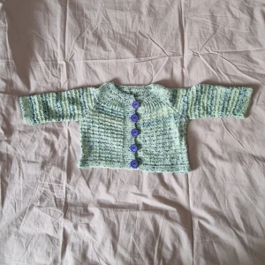 Stickad kofta med blå knappar - Säljer en söt stickad kofta i ljusgrön och blåmelerad färg med fem blå knappar framtill. Koftan har långärmad design och är gjord i mjukt stickat material, perfekt för kyliga dagar. Passar barn och har en klassisk rund halsringning. Det finns ingen storleksmärkning men jag skulle säga att den motsvarar stl 68 ca.  Ärmen är ca 15-16 cm från "armhålan" och den är ca 24 cm från halslinningen och ner.