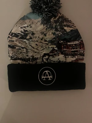 All cap 1 mössa  - Säljer en snygg mössa från All Cap1 med vinterlandskap och svart uppvikt kant med broderad logga. Mössan har en stor svartvit boll på toppen och kommer med en svart påse med logga. Perfekt accessoar för kalla dagar.