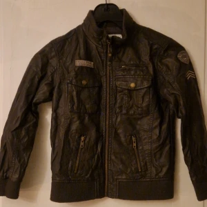 Mörkbrun skinnjacka från Vertbaudet - Säljer en mörkbrun jacka i fejkskinn från Vertbaudet med coola detaljer som bröstfickor med knappar, dragkedjor och patchar på ena ärmen. Jackan har ribbade muddar vid ärmslut och nederkant samt hög krage. Perfekt för den som vill ha en tuff look. Hund finns i hemmet. Kolla in mina andra annonser för samfrakt! 