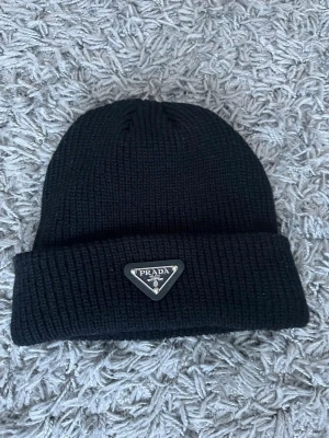 Svart stickad mössa från Prada - Säljer en svart stickad mössa från Prada med klassisk uppvikt kant och den ikoniska trekantiga metall-loggan framtill. Mössan är enkel och stilren, perfekt för dig som vill ha en clean look med exklusiv känsla.