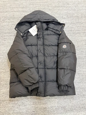 Svart Moncler jacka  - Svart pufferjacka från Moncler med huva och klassisk logga på ärmen. Jackan har quiltad design, elastiska muddar och tryckknappar framtill. Insidan har en cool serietidningsetikett och praktiska innerfickor. Perfekt för kalla dagar och riktigt snygg streetstil.